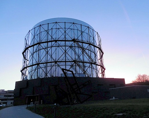 Zylinderförmiges, mit Fenstern verkleidetes Gasometer im Schein der untergehenden Sonne.