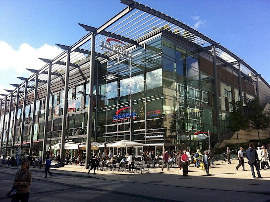 Gläserne Fassade der Schlössle-Galerie mit Fußgängern und Besuchern rund um den Eingang, die mit Cafés gesäumt ist.