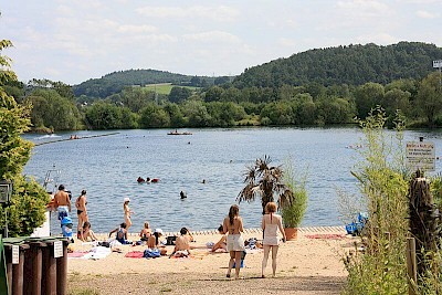 Sonnen beleuchteter Strandausschnitt des Niederweimarer Sees mit Palmen und Badegästen im Wasser sowie am Ufer.
